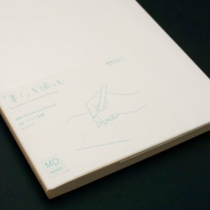 Peut inclure: Un carnet de notes blanc avec le texte "MD Notebook Journal A5 Dot Grid" et un dessin d'une main écrivant sur une page. Le texte "「書く」を愉しむ Designed for Comfortable Writing" est également sur la couverture.