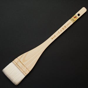 May include: A large, flat paintbrush with a long, light-colored wooden handle. The brush head features a thick, rectangular bundle of white bristles. Text is printed on the handle. A hole is drilled at the end of the handle.