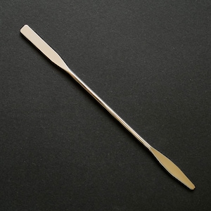 Può includere: Una spatola color argento con un lungo manico sottile e due estremità piatte a forma di paletta. La spatola è realizzata in metallo lucido ed è posta su uno sfondo scuro. È uno strumento utilizzato per mescolare o spalmare.