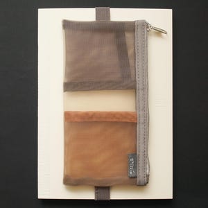Peut inclure: Un carnet beige avec un organiseur de poche en filet dans les tons marron et beige. L'organisateur a une fermeture éclair et une petite étiquette. Une sangle marron foncé fixe l'organisateur au carnet.