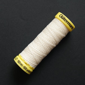 May include: A spool of cream-coloured thread with a yellow cap, branded "Gütermann" in black. The spool contains approximately 50-55 yards of thread. The thread is tightly wound, and the spool is set against a dark background.