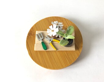 May include: A miniature wooden gardening set with a small rake, a pair of yellow clippers, and a white flower in a brown pot. The set is on a round wooden base.