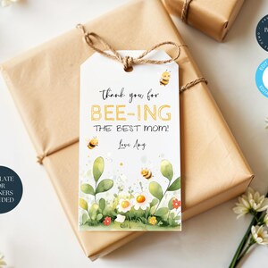 May include: A white gift tag with a bee theme. The tag features a watercolour illustration of bees, flowers, and leaves. The text on the tag reads "Thank you for bee-ing the best mom! Love Any".