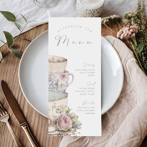 May include: A decorative afternoon tea menu card featuring stacked floral teacups and roses. The menu lists savoury and sweet options, such as ham & cheese, Victoria sponge, and scones. The card is on a white plate with gold cutlery.