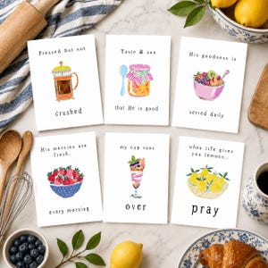 May include: Six white cards with watercolor illustrations and text. The cards feature images of a French press, jam, fruit bowl, strawberries, parfait, and lemons. Text includes phrases like "Pressed but not crushed" and "When life gives you lemons...pray."