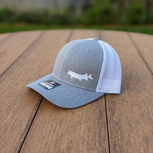 May include: A gray and white trucker hat with a white embroidered fish logo on the front. The hat has a gray front panel and bill, with a white mesh back. The hat is sitting on a wooden surface.