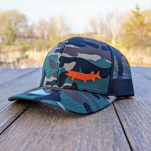 May include: Camouflage baseball cap with a black mesh back and a curved brim. The cap features an orange fish patch on the front. The hat is sitting on a wooden surface.