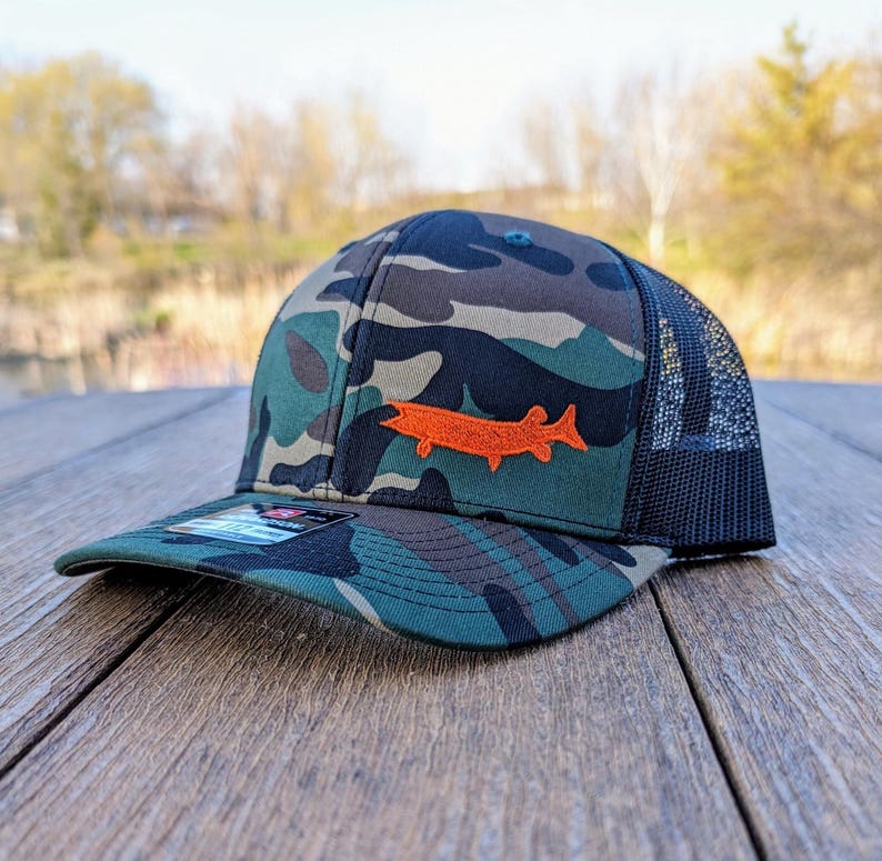 May include: Camouflage baseball cap with a black mesh back and a curved brim. The cap features an orange fish patch on the front. The hat is sitting on a wooden surface.