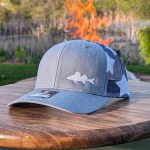 May include: A gray and black baseball cap with a white walleye fish logo on the front and a black and white star pattern on the back.