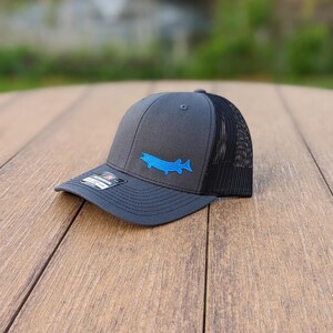 May include: A charcoal gray trucker hat with a black mesh back. The hat features a blue embroidered fish design on the front. The hat is sitting on a wooden surface.