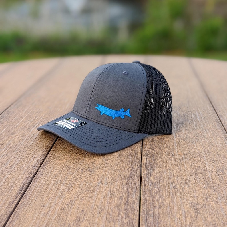 May include: A charcoal gray trucker hat with a black mesh back. The hat features a blue embroidered fish design on the front. The hat is sitting on a wooden surface.