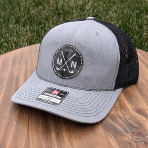 May include: A gray and black trucker hat with a black leather patch featuring a camouflage pattern, crossed golf clubs, and the letters "MN". The hat is adjustable and has a black mesh back.