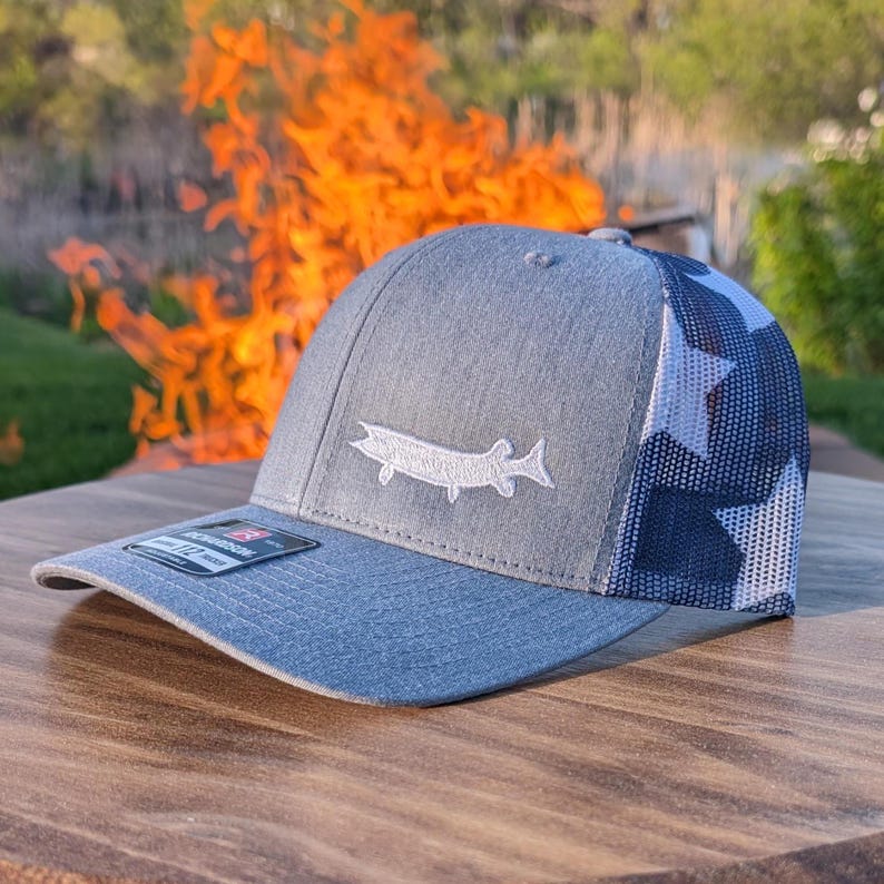 May include: A gray and blue baseball cap with a white fish emblem. The cap features a mesh back with a navy blue and white star pattern. The brim is blue, and the front is gray.