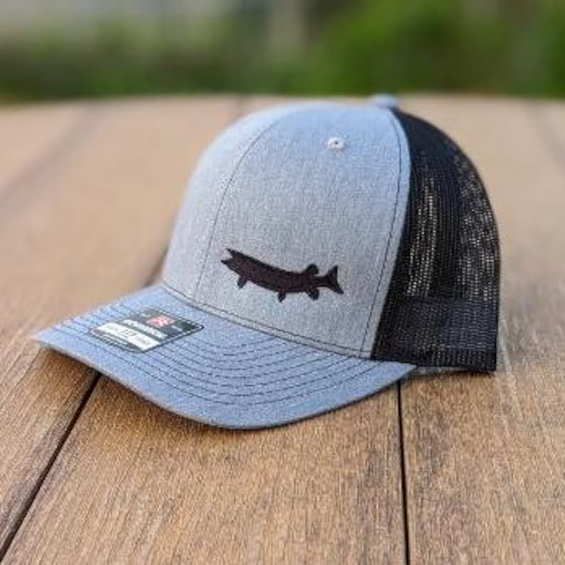 May include: A blue and black trucker hat with a fish silhouette embroidered on the front. The hat has a mesh back and a curved brim. The hat is sitting on a wooden surface.