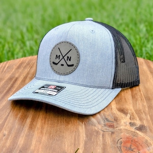 May include: A gray and black trucker hat with a gray leather patch featuring a hockey stick and puck design with the letters "MN".