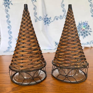 May include: Two decorative candle holders. Each has a woven, cone-shaped top in shades of brown and black, atop a clear glass base. The base is supported by a dark metal filigree stand. The candle holders are displayed on a wooden surface.
