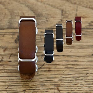 Puede incluir: Una colección de cinco correas de reloj de cuero con hebillas y trabillas de metal plateado. Las correas se presentan en varios colores, incluyendo marrón claro, negro, marrón oscuro, marrón medio y rojo intenso, con acabado liso.