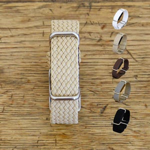 Op de afbeelding: Een beige geweven horlogeband met een textuurrijk vlechtpatroon en zilverkleurige metalen hardware. De band is voorzien van een centrale gesp en twee verschuifbare lussen. Aan de rechterkant zijn extra kleurvarianten in een kolom weergegeven.