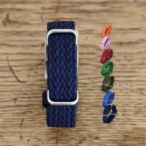 Könnte beinhalten: Ein dunkelblaues, geflochtenes Uhrenarmband mit einer silberfarbenen Metallschnalle. Rechts daneben zeigt eine vertikale Spalte sieben weitere Uhrenarmbänder in Orange, Hellrosa, Rot, Grün, Olivgrün, Hellblau und Dunkelblau, jeweils mit silberner Hardware.