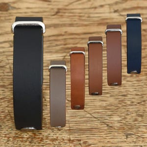 May include: A collection of six leather bracelets arranged in a diagonal line, featuring varying shades of black, gray, tan, brown, and navy blue. Each bracelet includes a silver-toned metal loop and a small circular metal stud fastener.
