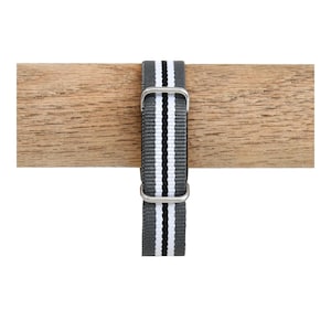 May include: A grey nylon watch strap featuring a central white stripe flanked by two thin black lines. The strap includes two silver-toned metal rectangular loops for adjustment and security, providing a functional and durable design for a timepiece.