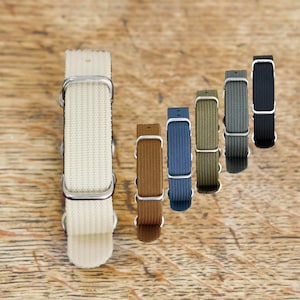 Op de afbeelding: Een collectie van zes nylon horlogebandjes met zilverkleurige metalen gespen en houders. De bandjes zijn in een rij gerangschikt en tonen een verscheidenheid aan kleuren, waaronder crème, bruin, blauw, olijfgroen, grijs en zwart, allemaal geweven.