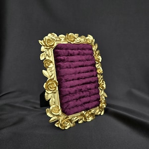 May include: A rectangular jewelry display stand featuring a deep purple velvet insert with horizontal slots for rings. The frame is finished in a metallic gold color and is decorated with a relief pattern of sculpted roses and leaves.