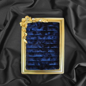 Puede incluir: Un expositor de joyas rectangular con marco dorado e interior de terciopelo azul oscuro. Un lazo dorado adorna la esquina superior izquierda. El estuche está diseñado para guardar anillos u otras joyas pequeñas.