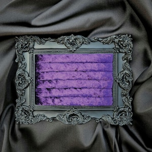 May include: A rectangular, ornate black frame with a plush, purple velvet interior. The frame has detailed scrollwork and is set against a dark, draped background, creating a luxurious display for jewelry or small items.