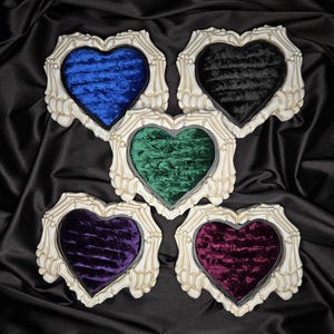 May include: Five heart-shaped decorative frames with a skeleton hand design. Each frame has a different colored velvet interior: blue, black, green, purple, and burgundy. The frames are off-white and sit on a black satin surface.