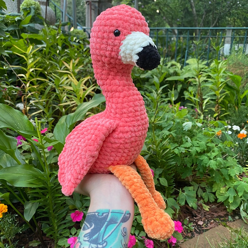 May include: A handmade, plush flamingo toy in shades of pink, white, black, and orange. The flamingo has a long neck, a black beak, and orange legs. The soft, textured material gives it a cuddly appearance, perfect for a child's room or nursery.