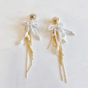 Op de afbeelding: Een paar witte parel- en gouden ketting oorhangers. De oorhangers hebben een cluster van witte parels in de vorm van een strik met gouden kettingkwastjes.