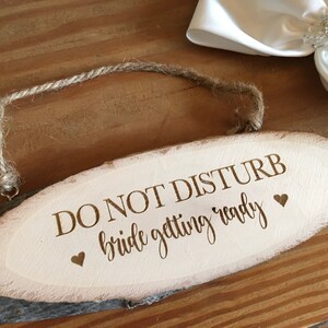 May include: Oval wooden sign with the text "DO NOT DISTURB bride getting ready" in a script font. The sign has a natural wood grain and a twine hanger. Two small heart symbols are on the sign.