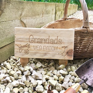 May include: A wooden sign with the text "Grandads Veg Patch" and a small illustration of a wheelbarrow and boots. The sign is sitting on a bed of small white stones.