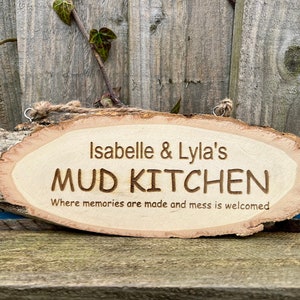 May include: A wooden sign with the text "Isabelle & Lyla's Mud Kitchen Where memories are made and mess is welcomed" carved into it.