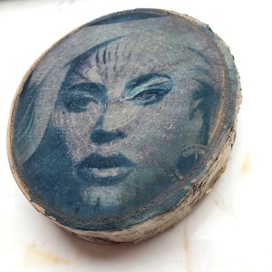 Op de afbeelding: Een close-up van een houten schijf met een blauwe en witte afbeelding van een vrouwen gezicht. De afbeelding is vervaagd en heeft een vintage look.