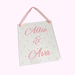 May include: White wooden sign with pink lettering that reads "Allie & Ava" with silver glitter dots scattered across the surface. The sign has a pink ribbon attached for hanging.