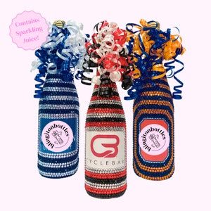 May include: Three sparkling juice bottles decorated with rhinestones and colorful ribbons. The bottles are decorated with blue and white stripes, red and black stripes, and orange and blue stripes. Each bottle has a round sticker with the text "blingitonbottles" and a small graphic of a bottle with a bow. The middle bottle has a sticker with the text "CYCLEBAR" and a red letter "G".