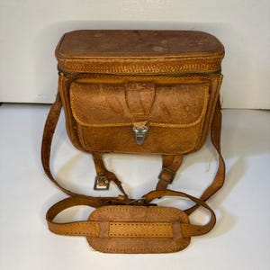 Puede incluir: Una bolsa de cámara vintage de cuero marrón con forma rectangular, un bolsillo frontal y una cremallera superior. La bolsa tiene una correa para el hombro y un asa, y un cierre metálico. El cuero muestra signos de desgaste.