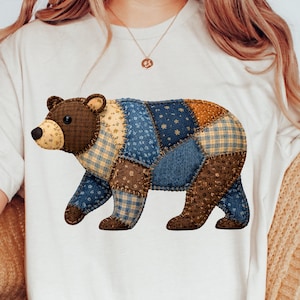 Könnte beinhalten: Ein weißes T-Shirt mit einem Patchwork-Bären-Design. Der Bär besteht aus verschiedenen Stoffmustern in Braun-, Blau- und Gelbtönen. Der Bär geht nach rechts. Das Shirt ist ein lässiges Kleidungsstück für den Alltag.