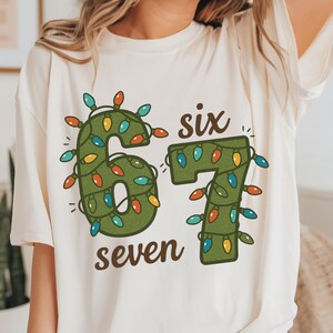 May include: Cream-colored t-shirt featuring a festive design. The number "67" is displayed in green, adorned with colorful Christmas lights. The words "six" and "seven" are written in brown, completing the holiday-themed graphic.