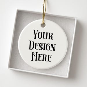 May include: A white, round ceramic ornament with the text "YOUR DESIGN HERE" in black. The ornament hangs from a gold string and is presented inside a white box.