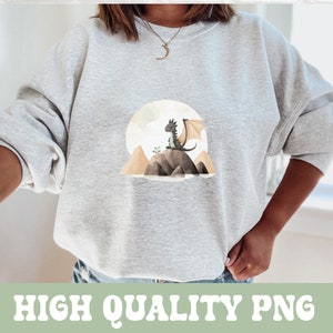 K&ouml;nnte beinhalten: Ein graues Sweatshirt mit einem Aufdruck eines kleinen, grauen Drachen, der auf einem Felsen mit einem Berg im Hintergrund sitzt. Der Drache hat einen hellgr&uuml;nen Fl&uuml;gel und blickt nach links.