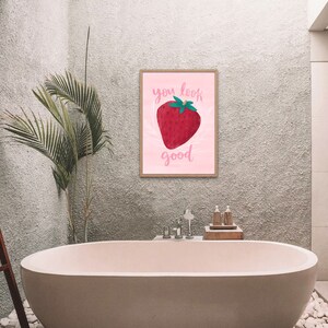 Painterly big strawberry graphic on a pink background with flourishes. Text is calligraphy with words You Look Good. Pun on You Look Berry Good. Art print is hanging above pink bathtub.