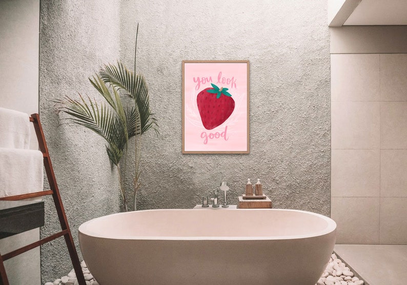 Painterly big strawberry graphic on a pink background with flourishes. Text is calligraphy with words You Look Good. Pun on You Look Berry Good. Art print is hanging above pink bathtub.