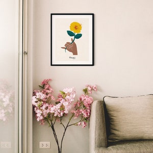 May include: A framed print of a yellow hibiscus flower being held by a hand making the Hawaiian shaka sign. The print has the word "Hawaii" written below the image.