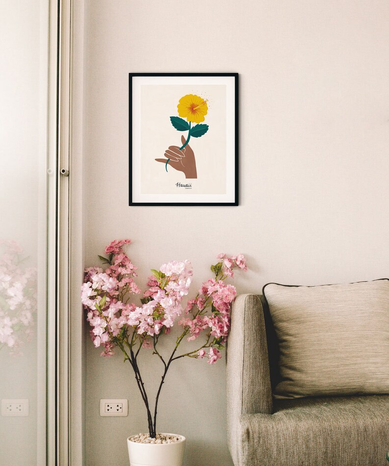 May include: A framed print of a yellow hibiscus flower being held by a hand making the Hawaiian shaka sign. The print has the word "Hawaii" written below the image.