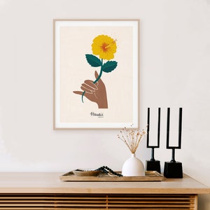 May include: A framed print of a yellow hibiscus flower with green leaves held in a brown hand with the word "Hawaii" written below the flower.