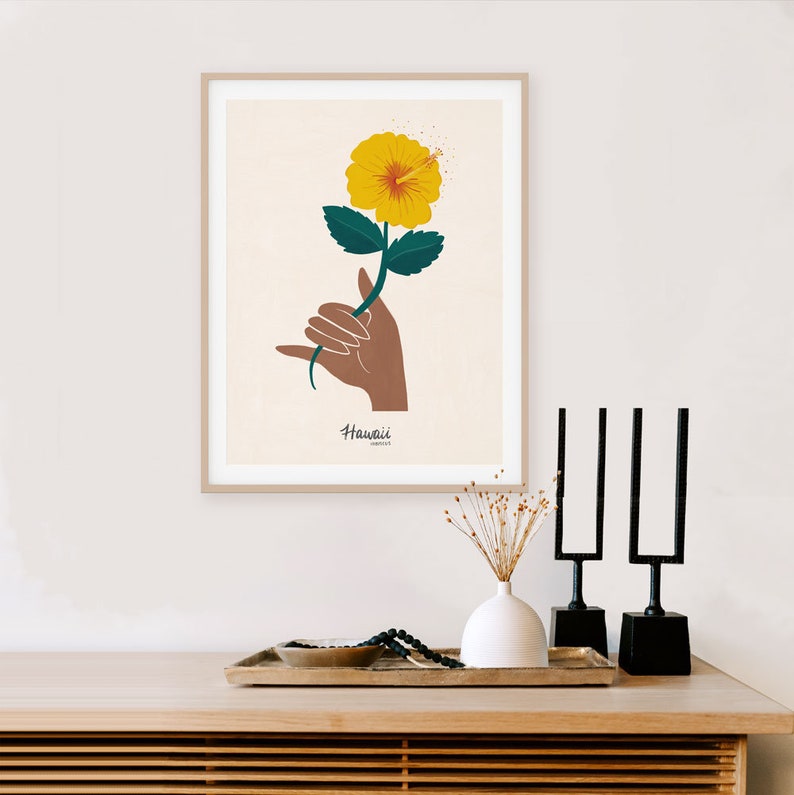 May include: A framed print of a yellow hibiscus flower with green leaves held in a brown hand with the word "Hawaii" written below the flower.