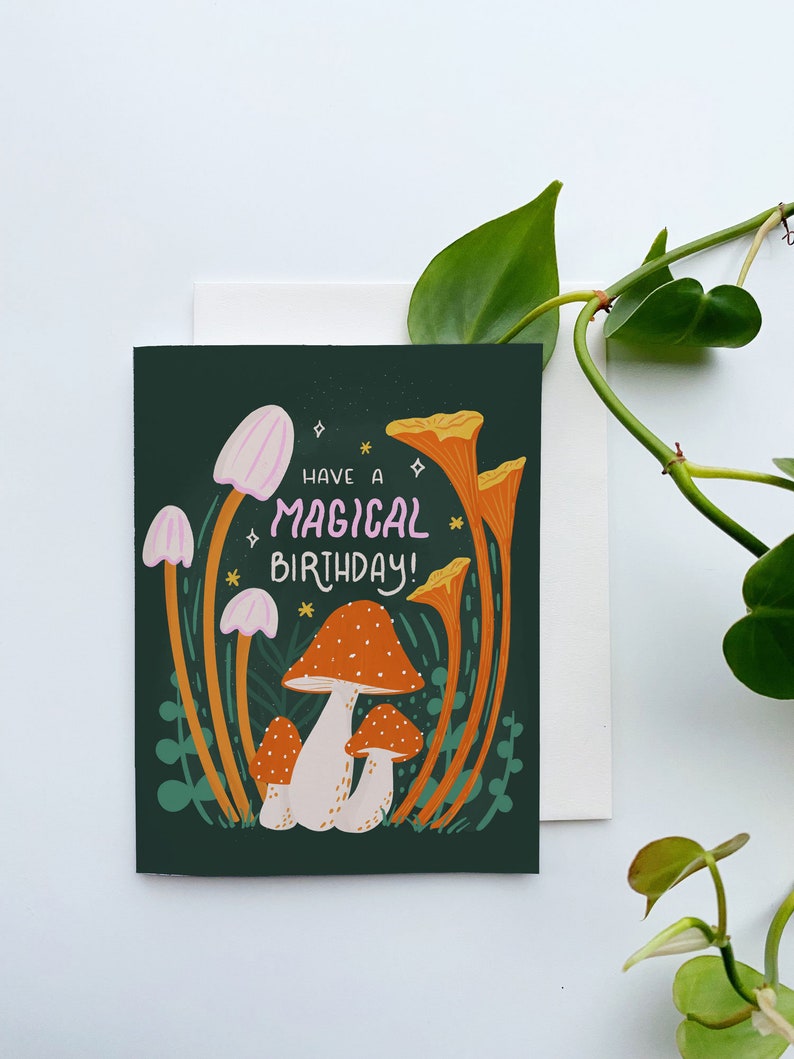 Greeting card with words "Have a magical birthday!" with trippy orange, beige, pink mushrooms (shiitake, enoki, toadstool, chanterelles) around with foliage and stars on a dark forest green background